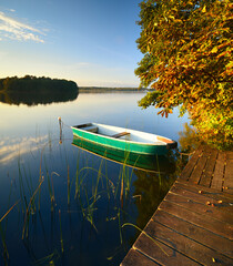 Horse Chestnut Tree by calm lake in full autumn colour at sunset and a rowing boat tied to a wooden pier