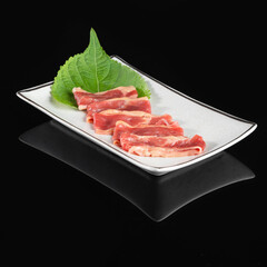 Thinly sliced raw meat arranged on a white plate with green leaf garnish, isolated on black background.