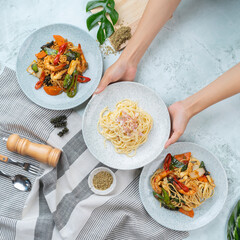 Three delicious pasta dishes served on gray plates with vegetables, viewed from above with a striped cloth and herbs.