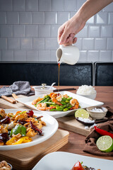 Hand pouring sauce over a delicious plate of vegetables and shrimp in a restaurant setting with other dishes in the background.