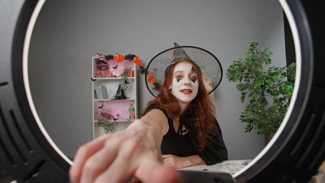 Young woman beauty blogger in witch costume at home films video tutorial for halloween party makeup on smartphone.
