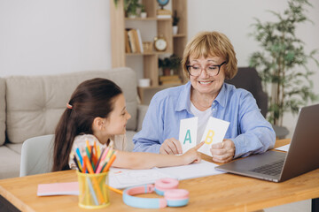 Fototapeta premium Happy grandma helping adorable preschool granddaughter school kid with homework task. Positive school pupil girl and granny reading learning book at laptop together, enjoying education at home