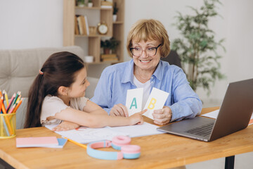 Happy grandma helping adorable preschool granddaughter school kid with homework task. Positive school pupil girl and granny reading learning book at laptop together, enjoying education at home