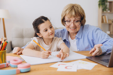 Happy grandma helping adorable preschool granddaughter school kid with homework task. Positive school pupil girl and granny reading learning book at laptop together, enjoying education at home