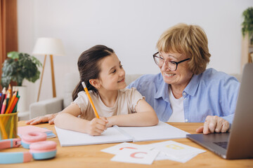 Happy grandma helping adorable preschool granddaughter school kid with homework task. Positive...