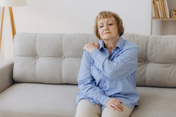 Senior woman sitting on sofa with shoulder pain. Elderly woman is enduring awful ache. Old woman suffering from shoulder injury, health problem concept