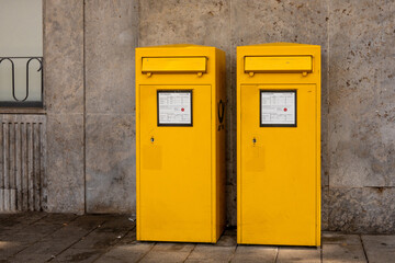 Two vibrant yellow mailboxes stand side by side against a gray stone wall