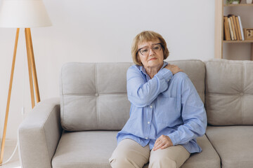 Obraz premium Senior woman sitting on sofa with shoulder pain. Elderly woman is enduring awful ache. Old woman suffering from shoulder injury, health problem concept