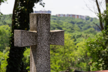 A close-up of a stone cross standing on an elevated hill, with a forest and city skyline in the background. The scene is bathed in sunlight on a bright summer day.
