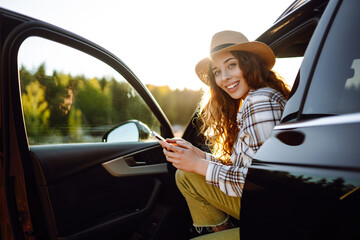 Young woman enjoying a sunny day by the lake while checking her phone next to a parked car in the...