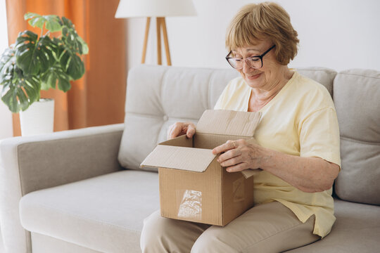 Happy senior woman sitting on couch opening carton box received parcel package, internet order, medical goods, pills, vitamins, medications, drugs.