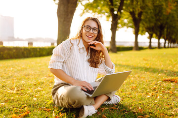 Young woman working on a laptop outdoors in a sunny park during the afternoon, enjoying nature and the warm weather. Online education, Freelance work, technology concept.