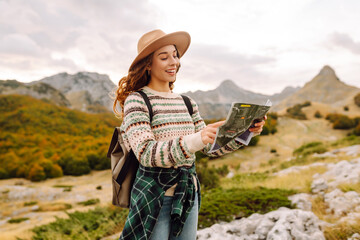 Young woman hiking in autumn landscape with a map, enjoying the scenic views of mountains and colorful trees in the background. Travel, nature concept