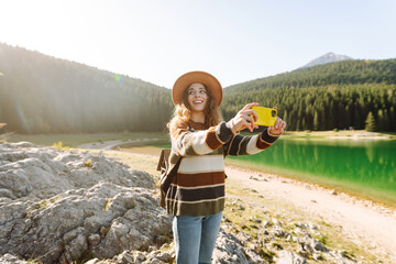 Young woman takes a photos the nice view by the serene lake surrounded by forested mountains during a sunny day in nature. Lifestyle, adventure, nature.