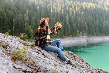 Young woman takes a photos the nice view by the serene lake surrounded by forested mountains during a sunny day in nature. Lifestyle, adventure, nature.