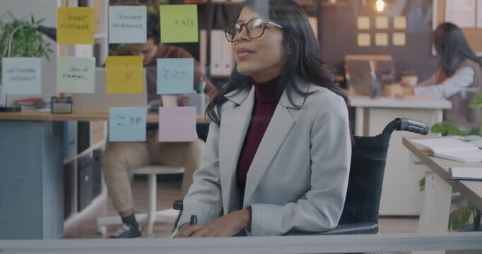 Creative young businesswoman in wheelchair working with sticky notes on glassboard in office focused on business development. Inclusive workspace and start-up concept.