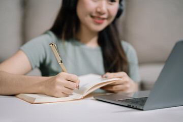 Smiling woman in headphones taking notes, motivated interested student studying online, using tablet, watching webinar training or listening to lecture, remote education concept.