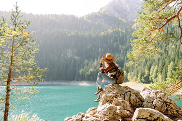 Traveler woman wearing a stylish hat and sweater enjoying a scenic lakeside landscape in the mountains during sunny autumn weather. Eco tourism. Active lifestyle.