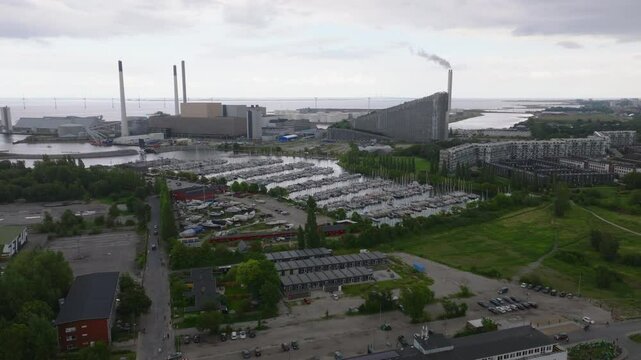 Aerial views capture Copenhagen's harbor, highlighting boats and the Amager Bakke or Copenhill waste to energy plant, which releases a plume of smoke into a cloudy summer sky