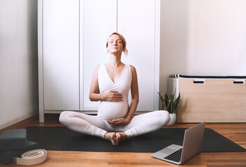 Pregnant woman practicing yoga online at home with laptop.