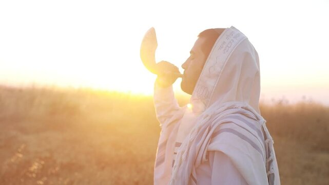 Jewish man in a traditional tallit prayer shawl blowing the ram's horn shofar, in the field against sunrise sky on Rosh HaShana and Yom Kippurim 