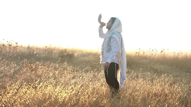 Jewish man in a traditional tallit prayer shawl blowing the ram's horn shofar, in the field against sunrise sky on Rosh HaShana and Yom Kippurim 
