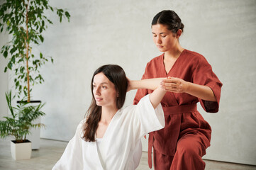 Masseuse making thai yoga massage. Woman therapist doing traditional massage treatment for female patient indoors. Client sitting, meditating, enjoying hand massage in calm atmosphere.