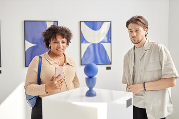Minimal waist up portrait of two young people looking at abstract art sculpture and wearing...