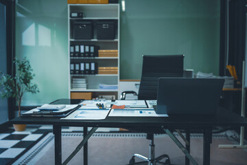 An empty office desk at night with a laptop displaying a business chart, balance sheet, loan calculator, graphs, and monthly budget stats. Blue and black tones, leather office chair.