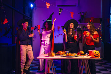 Asian friends, middle-aged man and young woman, enjoy Halloween office party with vampire costumes, fried chicken, grilled food, grape wine, champagne, RGB lights, festive decorations in the night.