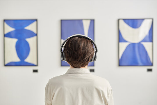 Minimal back view of unrecognizable man wearing headphones looking at abstract paintings in modern art gallery and listening to audio guide copy space
