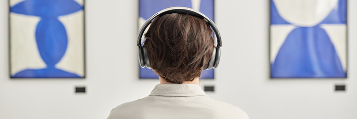 Header image with minimal back view of unrecognizable man wearing headphones looking at abstract paintings copy space