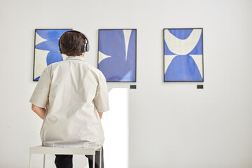 Back view of unrecognizable man looking at abstract art paintings and sitting on chair with headphones in minimal white museum setting copy space