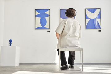 Full length back view of unrecognizable young man looking at abstract art paintings and sitting on chair while visiting modern art gallery lit by sunlight copy space