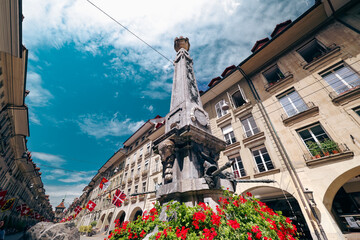 Fototapeta premium Historic fountain in Bern, Switzerland surrounded by blooming flowers and vibrant flags on a sunny day