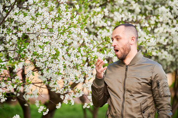 Man allergic suffering from seasonal allergy at spring in blossoming garden at springtime. Bearded young man sneezing and having runny nose in front of blooming tree. Spring allergy concept.