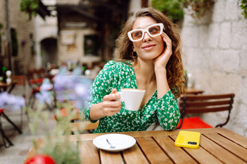 Woman enjoying coffee at an outdoor café in a charming town, dressed in a green floral dress during a sunny afternoon. People, fashion, lifestyle, travel and vacations