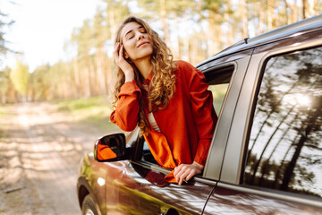 Naklejka premium The young woman leans out of the car window, basking in the sunlight and enjoying the natural surroundings of a serene forest road. The concept of active lifestyle, travel, tourism.