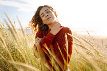 Young woman enjoying a sunny day outdoors in tall grass while wearing a cozy red sweater....