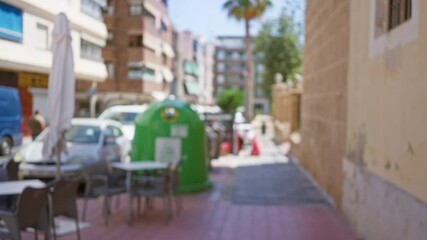 Blurry outdoor street scene with defocused buildings, parked cars, green recycling bin, and outdoor cafe in the foreground on a sunny day.