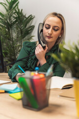 A woman in a green blouse is sitting at a desk, talking on a vintage telephone. She appears focused...