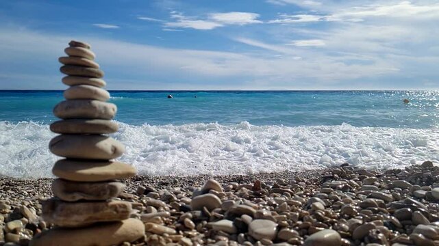 Stone Tower by the Blue Sea in Slow Motion a carefully balanced stack of smooth stones on a pebbled beach, with a bright blue sky and the ocean waves gently crashing in the background. Borsh, Alnania