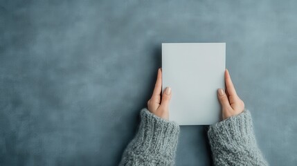 A pair of hands in a cozy grey sweater holds a blank square card against a blue background, symbolizing possibilities and invitations, waiting to be filled with personal messages.