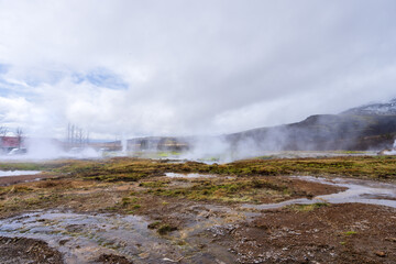 A misty geothermal landscape at Geysir, Iceland. Steam rises from numerous hot springs, creating a surreal and ethereal atmosphere. The volcanic activity is evident in the bubbling mud pools.