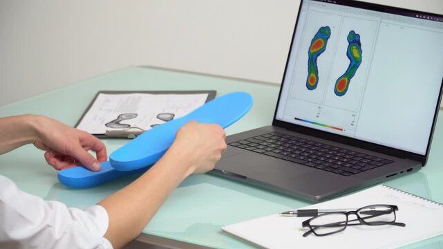 Female doctor orthopedist  inspecting custom made insoles in a clinic in front of the laptop with a test feet picture.