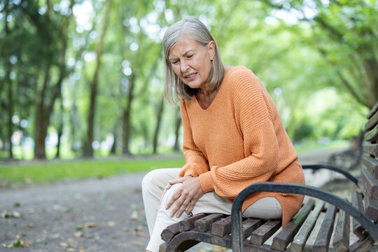 Senior woman feeling knee pain sitting on bench in park, showing discomfort. Mature adult dealing with joint pain outdoors in tranquil environment, highlighting aging and health challenges.