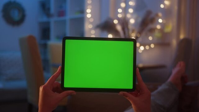 Hands holding a tablet with green screen display. Female relaxing on a sofa at home, Watching videos and reading social media or e-book. Close-up footage.