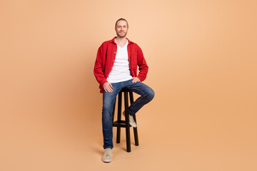 Full length portrait of nice young man sit stool wear red shirt isolated on beige color background