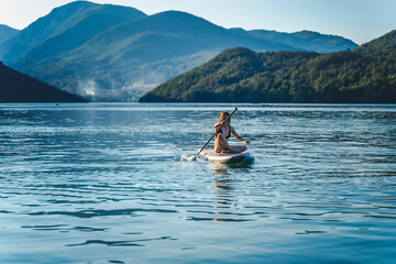 Young cheerful woman sitting with paddle on sup surf board on water in mountain lake