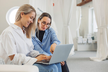 Two women collaborating on a laptop in a bright, modern workspace. One woman has long blonde hair and wears a white shirt, while the other has brown hair and glasses, dressed in a blue striped shirt.